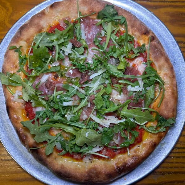A Celeste Restaurant Italian pizza with prosciutto, arugula, and shaved parmesan on a metal pan.