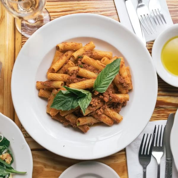 Overhead view of Celeste Restaurant's rigatoni bolognese with fresh basil, wine, and cutlery on a wooden table.