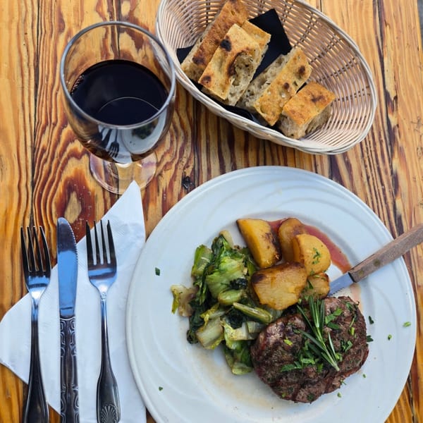Grilled steak with roasted potatoes, greens, red wine, and focaccia bread on a wooden table.