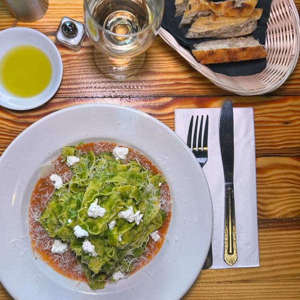 Overhead view of green pasta with ricotta and parmesan, bread, olive oil, and water on a wooden table.