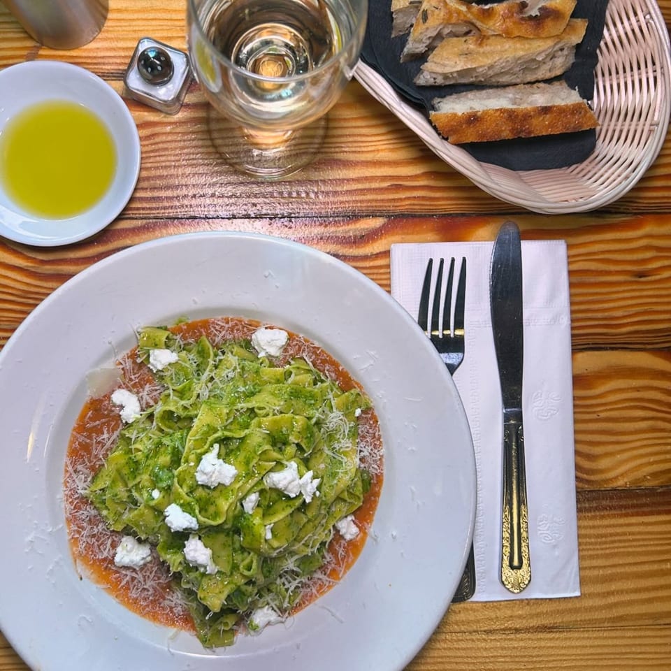 Overhead view of green pasta with ricotta and parmesan, bread, olive oil, and water on a wooden table.